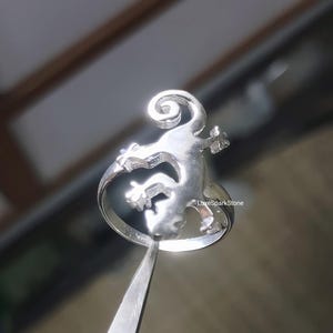 May include: A silver ring featuring a lizard design. The lizard is stylized with a curled tail and outstretched limbs. The ring is held by a pair of tweezers, showcasing the intricate details of the jewelry. The ring is a piece of jewelry.
