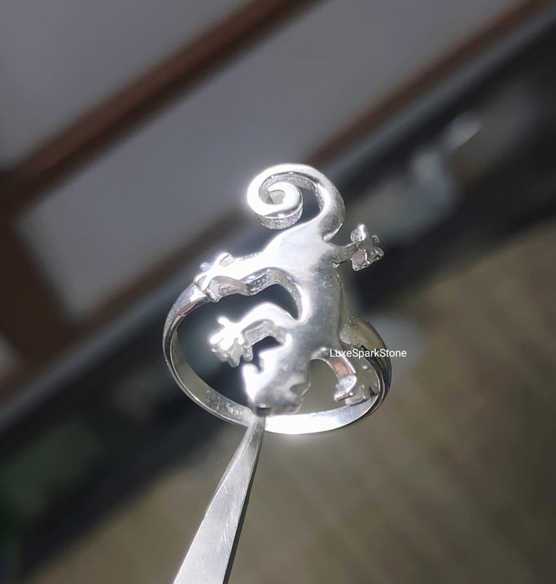 May include: A silver ring featuring a lizard design. The lizard is stylized with a curled tail and outstretched limbs. The ring is held by a pair of tweezers, showcasing the intricate details of the jewelry. The ring is a piece of jewelry.