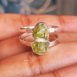 May include: Two sterling silver rings, each featuring a rough-cut, olive-green peridot gemstone. The rings are stacked, showcasing the natural texture and color of the stones. The rings are set against a warm, neutral background.