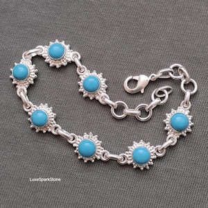 May include: A silver bracelet featuring turquoise-colored stones set in a sunburst design. The bracelet has a lobster clasp closure and is displayed on a gray surface. The bracelet is a piece of jewelry.