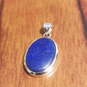 May include: Oval-shaped, deep blue pendant with a silver-coloured metal frame and bail. The pendant features a polished, speckled blue stone. The background is a warm, wooden surface.