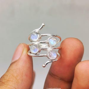 Könnte beinhalten: Ein silberner Bypass-Ring mit vier runden, irisierenden Mondsteinen in polierter Metallfassung. Die Ringschiene hat ein spiralförmiges Design, das sich um den Finger windet, wobei jedes Ende in einer kleinen, abgerundeten Silberkugel endet. Text LuxeSparkStone.