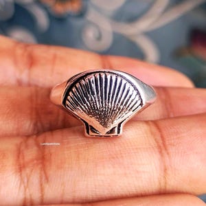 Könnte beinhalten: Ein silberner Ring mit einem detaillierten Muscheldesign. Der Ring hat ein glattes Band und einen strukturierten Muschelmittelteil. Der Ring wird auf einer Hand gezeigt.