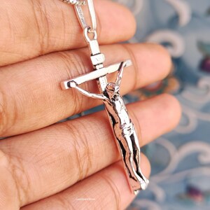 Puede incluir: Un colgante de crucifijo plateado con una representaci&oacute;n detallada de Jes&uacute;s en la cruz. El colgante cuelga de una delicada cadena de plata. La imagen est&aacute; sobre un fondo suave y borroso.
