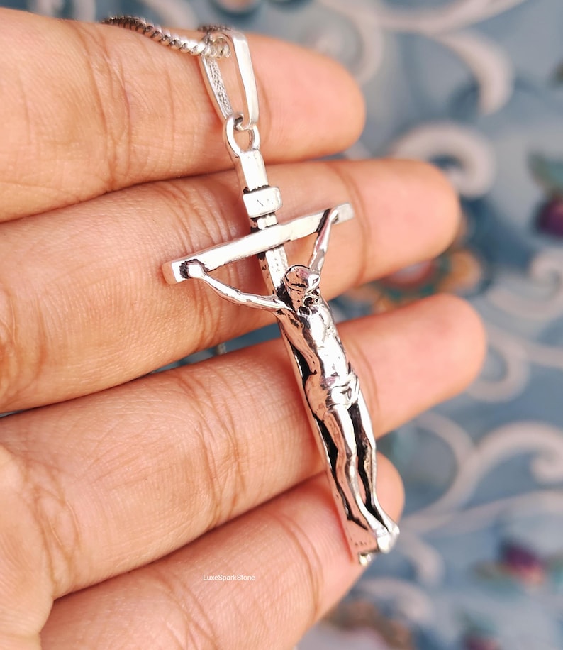 Puede incluir: Un colgante de crucifijo plateado con una representaci&oacute;n detallada de Jes&uacute;s en la cruz. El colgante cuelga de una delicada cadena de plata. La imagen est&aacute; sobre un fondo suave y borroso.