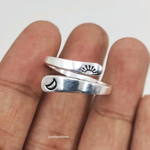 May include: A silver-toned bypass ring featuring an open wrap design. One end of the band displays a small engraved sun icon, while the other end features a crescent moon icon. The metal has a smooth, polished finish with dark etched details.