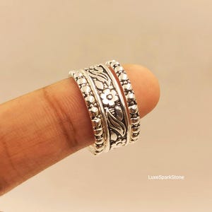 May include: A silver ring featuring a central band with an embossed floral and leaf pattern, flanked by two thinner bands with a beaded texture. The ring has a polished metallic finish and the text LuxeSparkStone is visible in the bottom right corner.