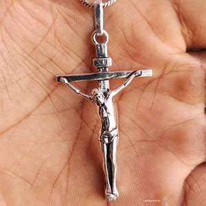 Puede incluir: Un colgante de crucifijo de plata con una detallada representaci&oacute;n de Jesucristo en la cruz. El colgante cuelga de una delicada cadena de plata. La cruz mide aproximadamente 5 cm de largo. El colgante se sostiene en una mano.