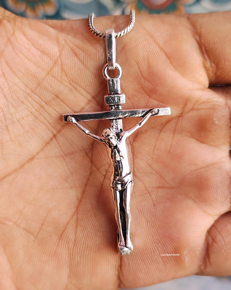 Puede incluir: Un colgante de crucifijo de plata con una detallada representaci&oacute;n de Jesucristo en la cruz. El colgante cuelga de una delicada cadena de plata. La cruz mide aproximadamente 5 cm de largo. El colgante se sostiene en una mano.