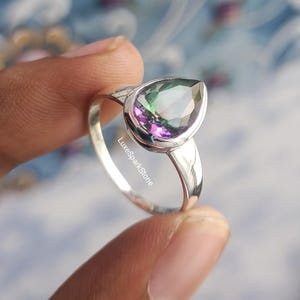 May include: A silver ring featuring a pear-shaped, iridescent gemstone with hues of green, purple, and blue. The ring's band is a simple, polished silver design. The text "LuxeSparkStone" is visible on the ring.