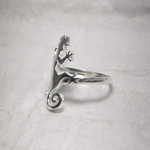 May include: A silver ring featuring a lizard design. The lizard is depicted in a side profile, with its tail curled and its legs extended. The ring is stamped with "LuxeSparkStone".