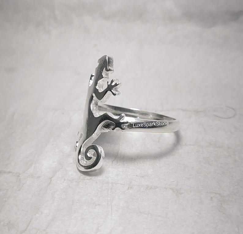 May include: A silver ring featuring a lizard design. The lizard is depicted in a side profile, with its tail curled and its legs extended. The ring is stamped with "LuxeSparkStone".