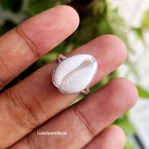 Può includere: Un anello in argento con una conchiglia di ciprea bianca come elemento centrale. La conchiglia ha una forma ovale liscia con una fessura centrale e bordi scanalati. La fascia in metallo è sottile e lucida, fissando la conchiglia in una montatura. Testo: LuxeSparkStone.