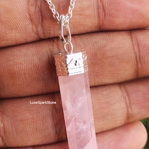 May include: A silver necklace featuring a pink rose quartz crystal pendant. The crystal is rectangular with a pointed base. The pendant hangs from a silver chain. The text "LuxeSparkStone" is visible on the crystal.