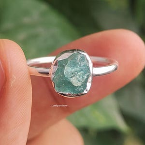 Könnte beinhalten: Ein Silberring mit einem rohen, oval geformten, hellblau-grünen Edelstein. Der Ring wird vor einem verschwommenen grünen Hintergrund gehalten. Der Edelstein hat ein strukturiertes, natürliches Aussehen.