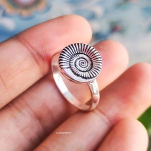 Könnte beinhalten: Ein silberner Ring mit einem kreisförmigen Spiraldesign. Der Ring hat eine detaillierte, strukturierte Scheibe mit einem zentralen Spiralmuster. Das Band ist glatt und poliert. Der Ring ist ein Schmuckstück.