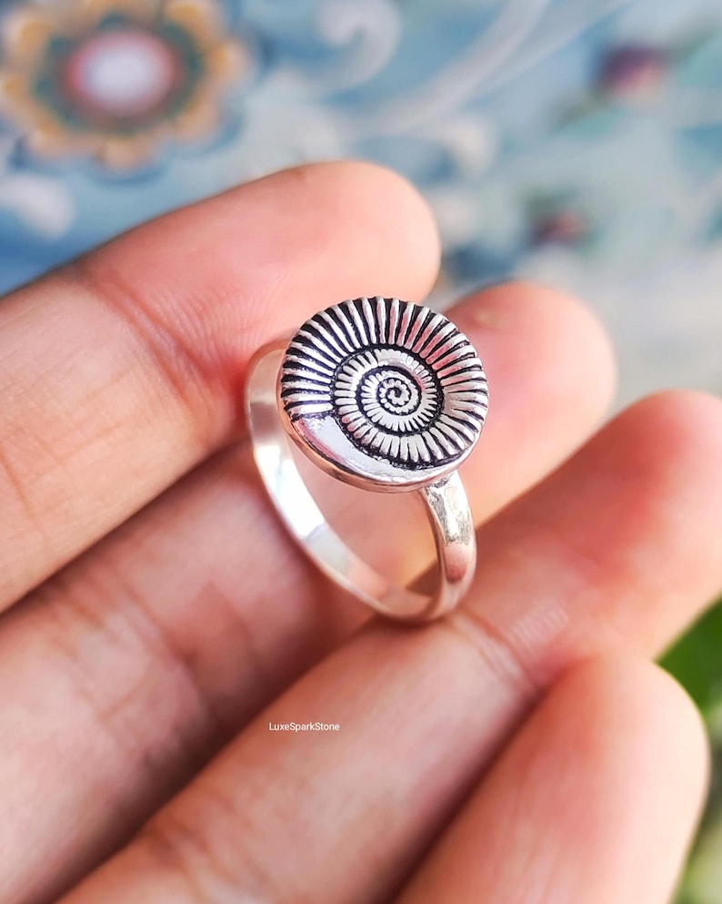 Pu&ograve; includere: Un anello d'argento con un design circolare a spirale. Il punto focale dell'anello &egrave; un disco strutturato e dettagliato con un motivo a spirale centrale. La fascia &egrave; liscia e lucida. L'anello &egrave; un gioiello.