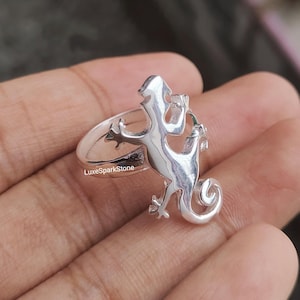 May include: A silver-colored ring featuring a detailed lizard design. The lizard is depicted with intricate features, including its legs, tail, and head. The ring has a smooth, polished finish and is likely made of sterling silver.