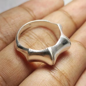 Könnte beinhalten: Ein silberner Ring mit einem Bambus-inspirierten Design. Der Ring hat eine Reihe von gebogenen Segmenten, die einen einzigartigen und organischen Look erzeugen. Das polierte Metall reflektiert das Licht und hebt die Details des Rings hervor. Eine Nahaufnahme zeigt die Handwerkskunst des Rings.