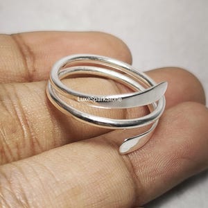 Könnte beinhalten: Ein silberfarbener Wickelring mit Spiraldesign. Der Ring hat eine glatte, polierte Oberfläche und ein mehrschichtiges, offenes Design. Der Ring wird auf einer Hand gezeigt.