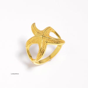 May include: A gold starfish ring. The ring has a detailed starfish design with textured surfaces and a smooth, rounded band. The ring is a statement piece, ideal for those who love ocean-themed jewellery. The ring is a statement piece.