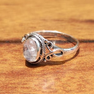 Pu&ograve; includere: Un anello d'argento con una gemma ovale e trasparente. L'anello presenta un design ornato attorno alla gemma e sulla fascia, con volute e dettagli testurizzati. Appoggiato su una superficie di legno calda.