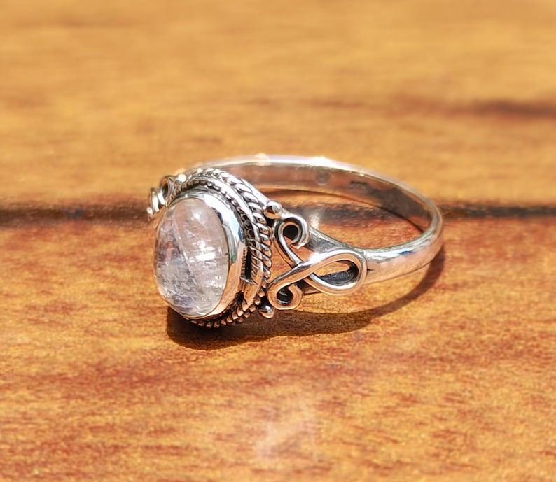 Pu&ograve; includere: Un anello d'argento con una gemma ovale e trasparente. L'anello presenta un design ornato attorno alla gemma e sulla fascia, con volute e dettagli testurizzati. Appoggiato su una superficie di legno calda.