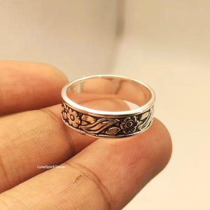 Könnte beinhalten: Ein silberner Ring mit floralem Design. Der Ring hat ein Band mit detaillierten Schnitzereien von Blumen und Blättern. Der Ring wird von den Fingern einer Person gehalten. Der Markenname "LuxeSparkStone" ist unten links sichtbar.