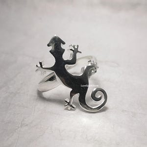 May include: A silver ring featuring a lizard design. The lizard is silhouetted, with a curled tail and outstretched limbs. The ring has a polished finish and a band that wraps around the finger.