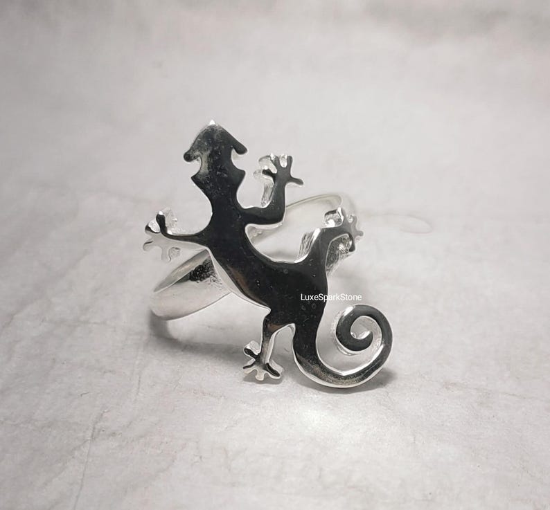 May include: A silver ring featuring a lizard design. The lizard is silhouetted, with a curled tail and outstretched limbs. The ring has a polished finish and a band that wraps around the finger.