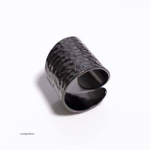 May include: A black, adjustable cuff ring with a hammered texture. The ring has a wide band and an open design, allowing for size adjustment. The surface has a dimpled pattern, creating a unique visual effect. The ring is made of a dark, possibly metal material.