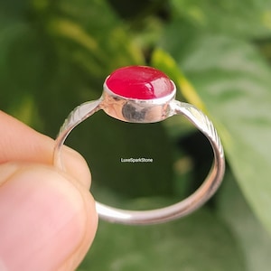 Könnte beinhalten: Ein silberner Ring mit einem leuchtend roten Edelstein. Der Ring hat einen glatten, oval geformten Stein, der in einem polierten Silberband gefasst ist. Das Band hat ein strukturiertes Design an den Seiten. Der Text "LuxeSparkStone" ist sichtbar.