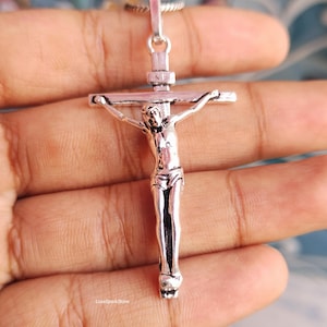 Puede incluir: Colgante de crucifijo plateado que representa a Jesucristo en la cruz, suspendido de una fina cadena. La imagen en primer plano revela los detalles de la joyer&iacute;a religiosa. S&iacute;mbolo cristiano tradicional.