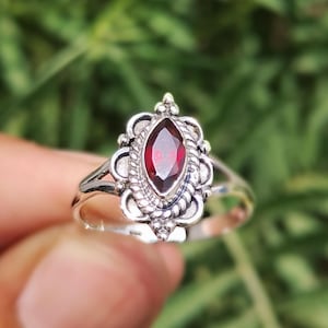 May include: A silver ring featuring a marquise-cut, deep red gemstone. The gemstone is set in an ornate, filigree-style mount. The ring's band splits into two delicate strands on either side of the gemstone.