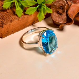 May include: A silver ring with a large, oval-cut, light blue gemstone. The gemstone is held in place by four prongs. The ring is shown on a light surface, with green leaves and brown wood in the background.