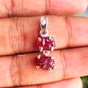 May include: A silver pendant with two raw, deep red ruby gemstones. The gemstones are irregularly shaped and set in silver. The pendant has a loop at the top for a chain. The design is simple and elegant. The pendant is likely to be worn as jewellery.