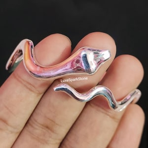 May include: Two silver snake rings. The rings are in a serpentine shape, with a head and a curved body. The rings are on a hand, with a dark background. The text "LuxeSparkStone" is visible.