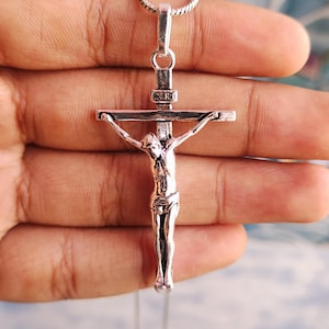 Puede incluir: Un colgante de crucifijo plateado con una representaci&oacute;n detallada de Jes&uacute;s en la cruz. El colgante cuelga de una delicada cadena. La imagen es un primer plano, que destaca los intrincados detalles de la joyer&iacute;a religiosa.
