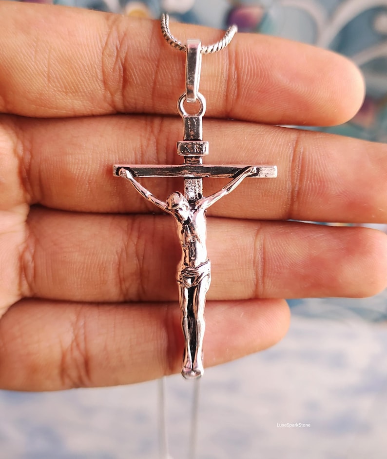 Puede incluir: Un colgante de crucifijo plateado con una representaci&oacute;n detallada de Jes&uacute;s en la cruz. El colgante cuelga de una delicada cadena. La imagen es un primer plano, que destaca los intrincados detalles de la joyer&iacute;a religiosa.