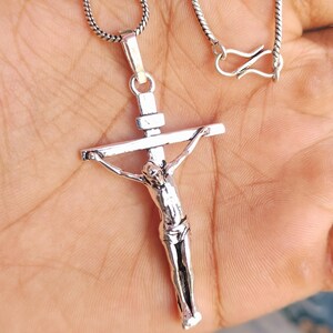 Puede incluir: Un colgante de crucifijo plateado en una cadena. El colgante presenta una detallada representaci&oacute;n de Jesucristo en la cruz. La cadena tiene un dise&ntilde;o texturizado y un cierre de cierre. El colgante se sostiene sobre un fondo color piel.