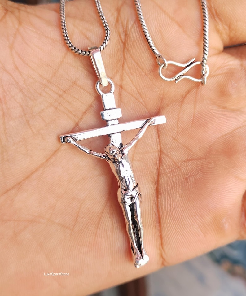Puede incluir: Un colgante de crucifijo plateado en una cadena. El colgante presenta una detallada representaci&oacute;n de Jesucristo en la cruz. La cadena tiene un dise&ntilde;o texturizado y un cierre de cierre. El colgante se sostiene sobre un fondo color piel.