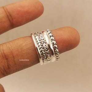 Könnte beinhalten: Eine Sammlung von Silberringen mit verschiedenen Designs, darunter strukturierte Muster und glatte Bänder. Die Ringe sind aufeinander gestapelt und zeigen die verschiedenen Stile und Texturen. Die Ringe werden auf einem Finger präsentiert.