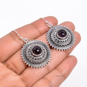 Könnte beinhalten: Ein Paar silberfarbene Ohrringe mit kreisförmigem Design. Jeder Ohrring hat einen zentralen, runden, dunkelvioletten Edelstein, der von aufwendigen Metallarbeiten umgeben ist. Die Ohrringe haben einen Hakenverschluss.