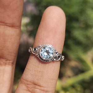 Peut inclure: Une bague en argent ornée d'une pierre précieuse ronde bleu clair. La bague présente un design détaillé et orné avec une bordure torsadée en forme de corde autour de la pierre et des volutes décoratives. La bague est sur un doigt.