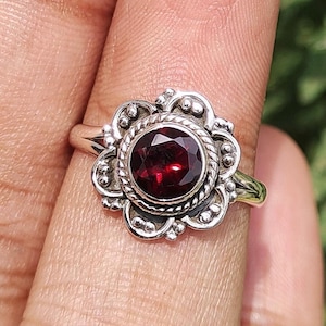 以下が含まれることがあります： 大きな丸い深紅色の宝石をあしらった、フローラルデザインのシルバーリング。 リングは、詳細なテクスチャード加工のバンドと装飾的なベゼルセッティングを備え、ヴィンテージまたはアンティークな美しさを生み出しています。