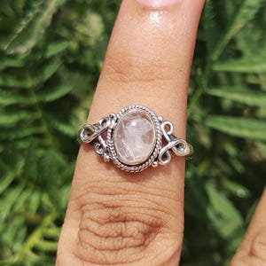 Pu&ograve; includere: Un anello d'argento con una pietra preziosa ovale rosa chiaro. L'anello ha un design ornato con un bordo a corda intrecciata attorno alla pietra e volute decorative sui lati. L'anello &egrave; mostrato su un dito.