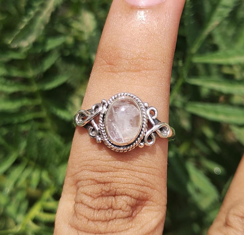 Pu&ograve; includere: Un anello d'argento con una pietra preziosa ovale rosa chiaro. L'anello ha un design ornato con un bordo a corda intrecciata attorno alla pietra e volute decorative sui lati. L'anello &egrave; mostrato su un dito.