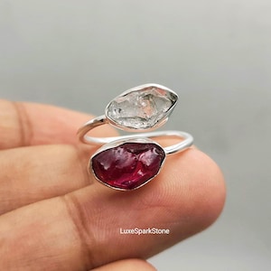 Könnte beinhalten: Ein offener Ring mit einem Silberband und zwei rohen, ungeschliffenen Edelsteinen. Ein Stein ist ein klarer, durchscheinender Kristall, der andere ist ein tiefroter, undurchsichtiger Stein. Der Text LuxeSparkStone ist unten im Bild zu sehen.