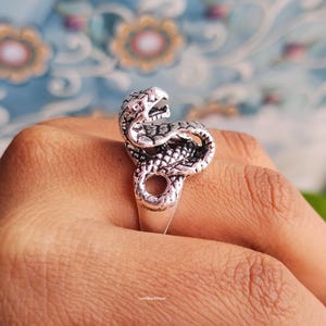 May include: A silver-colored ring in the shape of a snake, with detailed scales and an open mouth. The snake is coiled around the finger, creating a unique and eye-catching design. The ring is shown on a hand.