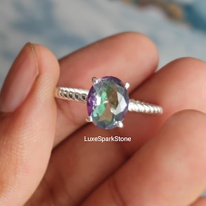 Peut inclure: Une bague en argent ornée d'une grosse pierre précieuse ovale. La pierre affiche un mélange de couleurs, notamment le violet, le vert et le bleu. Le bracelet présente un motif torsadé. Le texte "LuxeSparkStone" est visible.